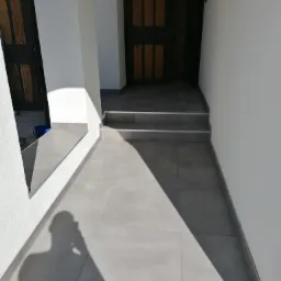 Jasnoszare, kwadratowe płytki podłogowe prowadzące do wejścia z dwoma stopniami, widoczne drzwi wejściowe i cień fotografa.