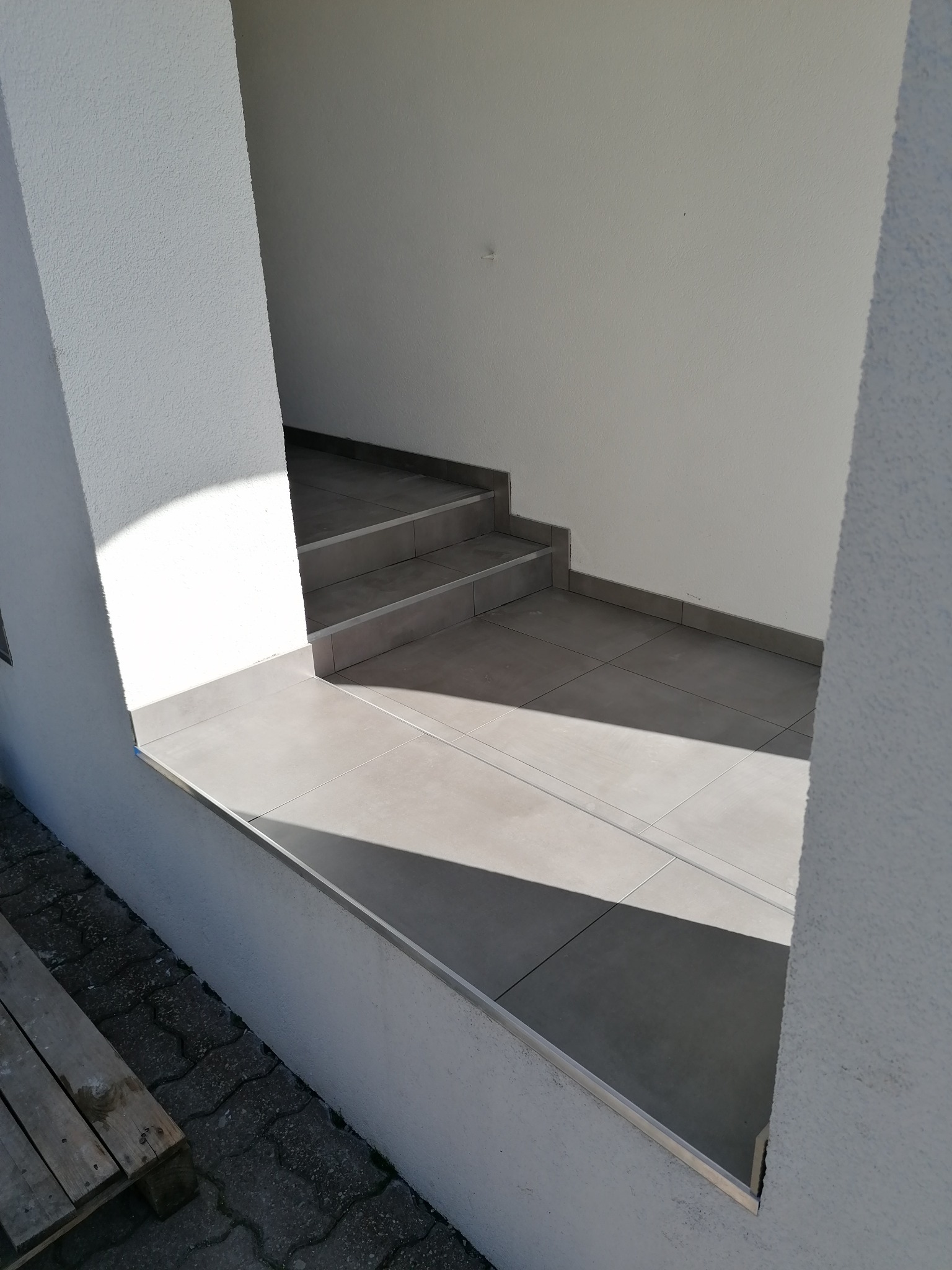 Wejście do budynku z trzema stopniami wyłożonymi szarą, kwadratową płytką ceramiczną, z widocznym profilem wykończeniowym na krawędziach i fragmentem drewnianej palety obok.