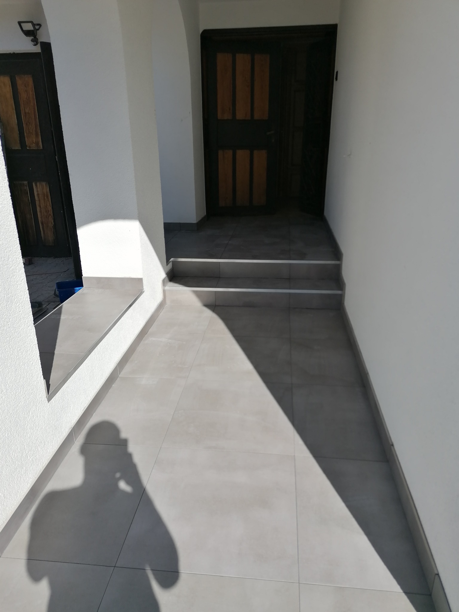 Jasnoszare, kwadratowe płytki podłogowe prowadzące do wejścia z dwoma stopniami, widoczne drzwi wejściowe i cień fotografa.