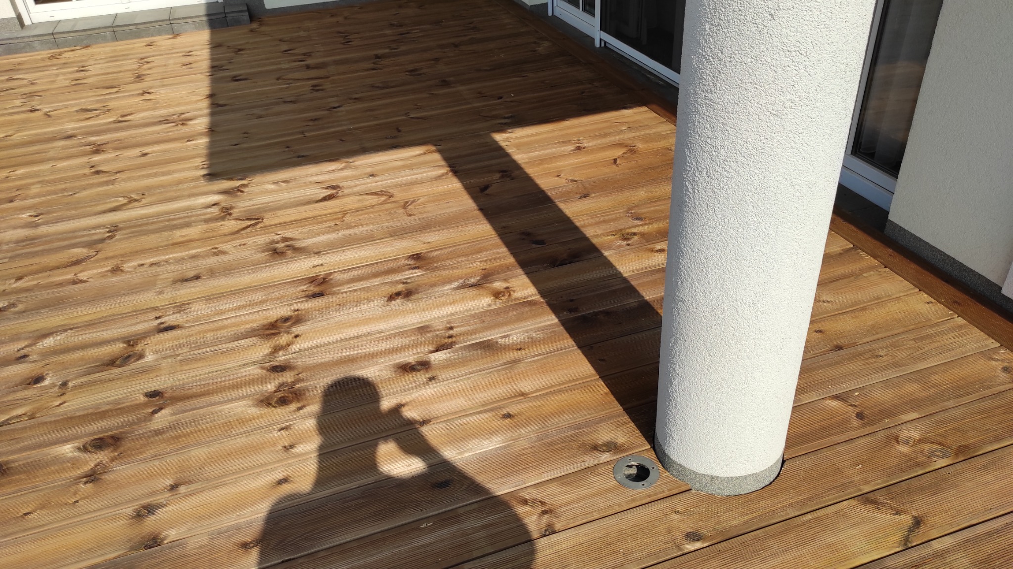 Drewniany taras z widocznym cieniem osoby robiącej zdjęcie, oświetlony słońcem, z filarem przy ścianie budynku i wbudowanymi punktami świetlnymi w podłodze.