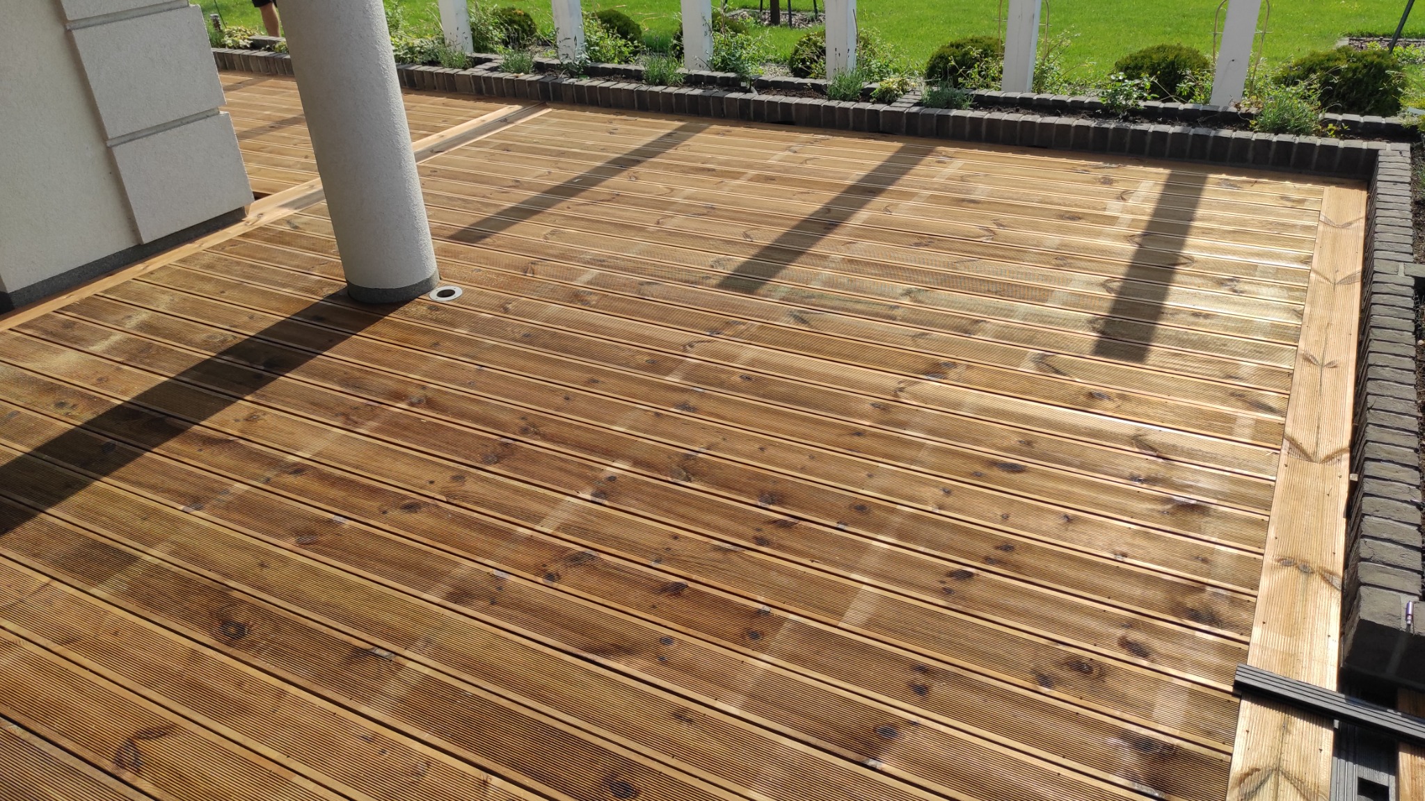 Nowa drewniana podłoga tarasowa z desek kompozytowych w kolorze jasnego drewna, z widocznym słupem podtrzymującym zadaszenie oraz obrzeżem z cegły klinkierowej i zielenią w tle.