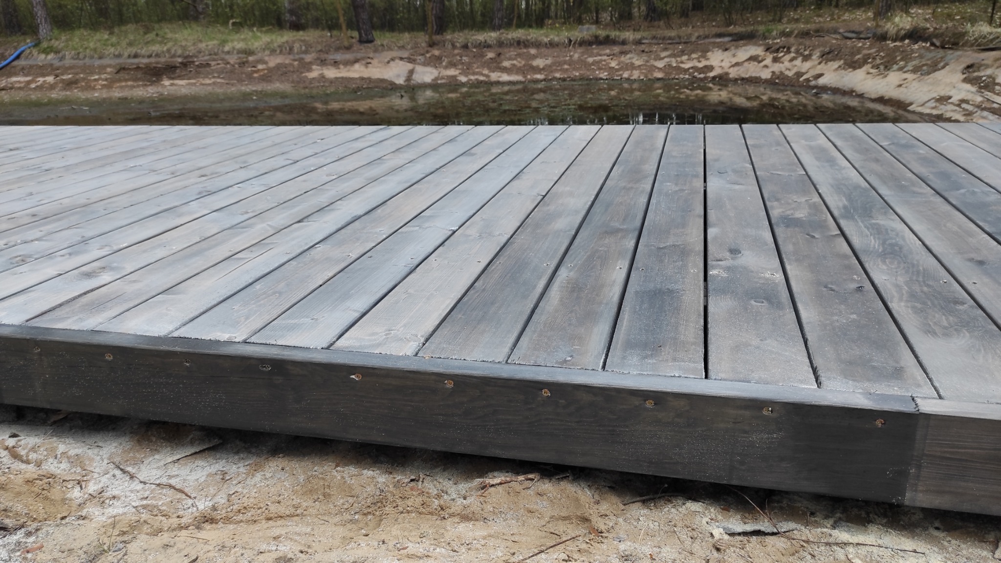 Drewniany pomost z desek w odcieniach szarości, widoczne wkręty mocujące, tło stanowi brzeg zbiornika wodnego i las.