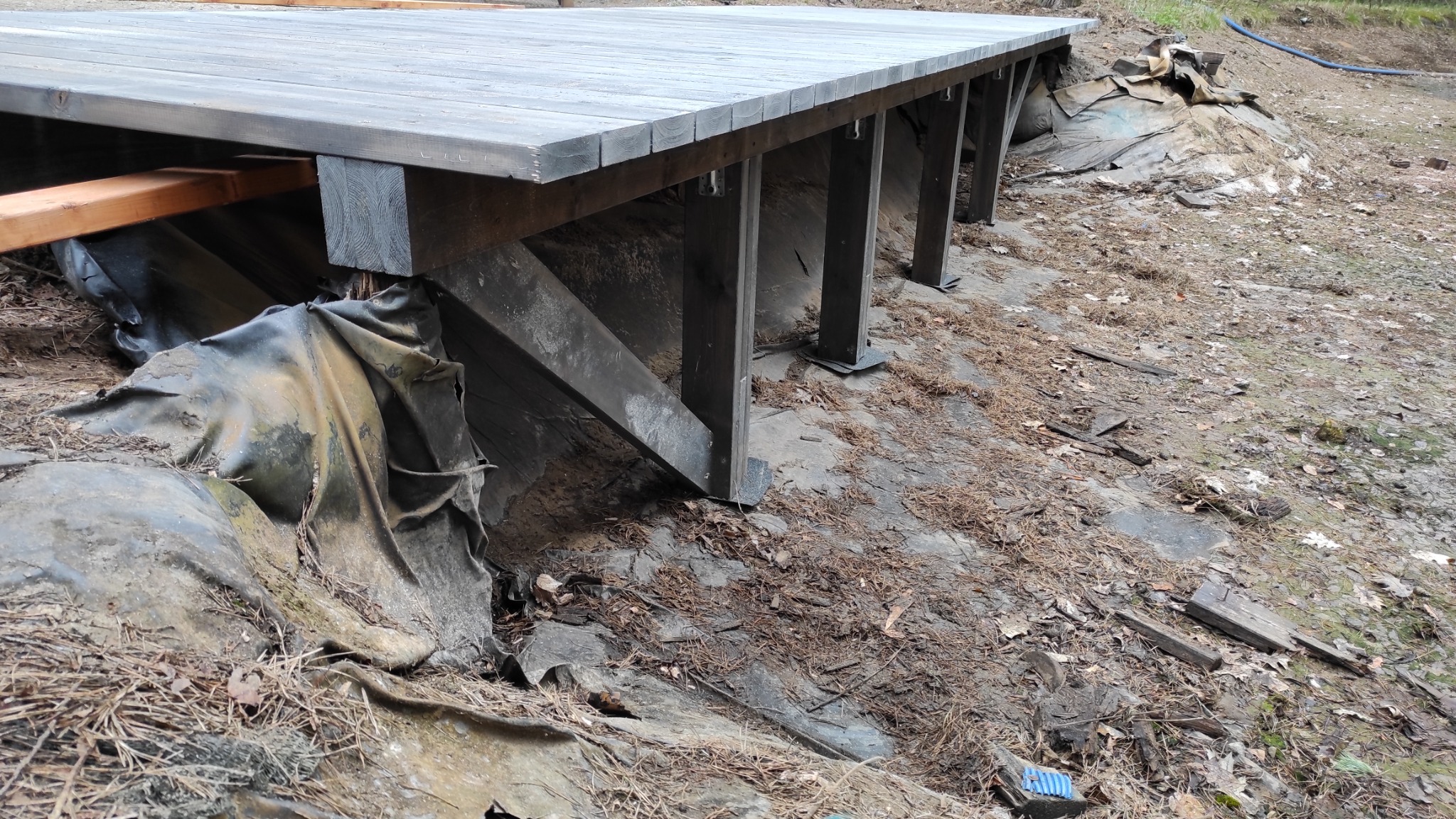 Drewniany podest na nierównym terenie, wsparty na ciemnych, drewnianych słupach z ukośnymi wzmocnieniami, widoczne zabezpieczenie terenu folią.