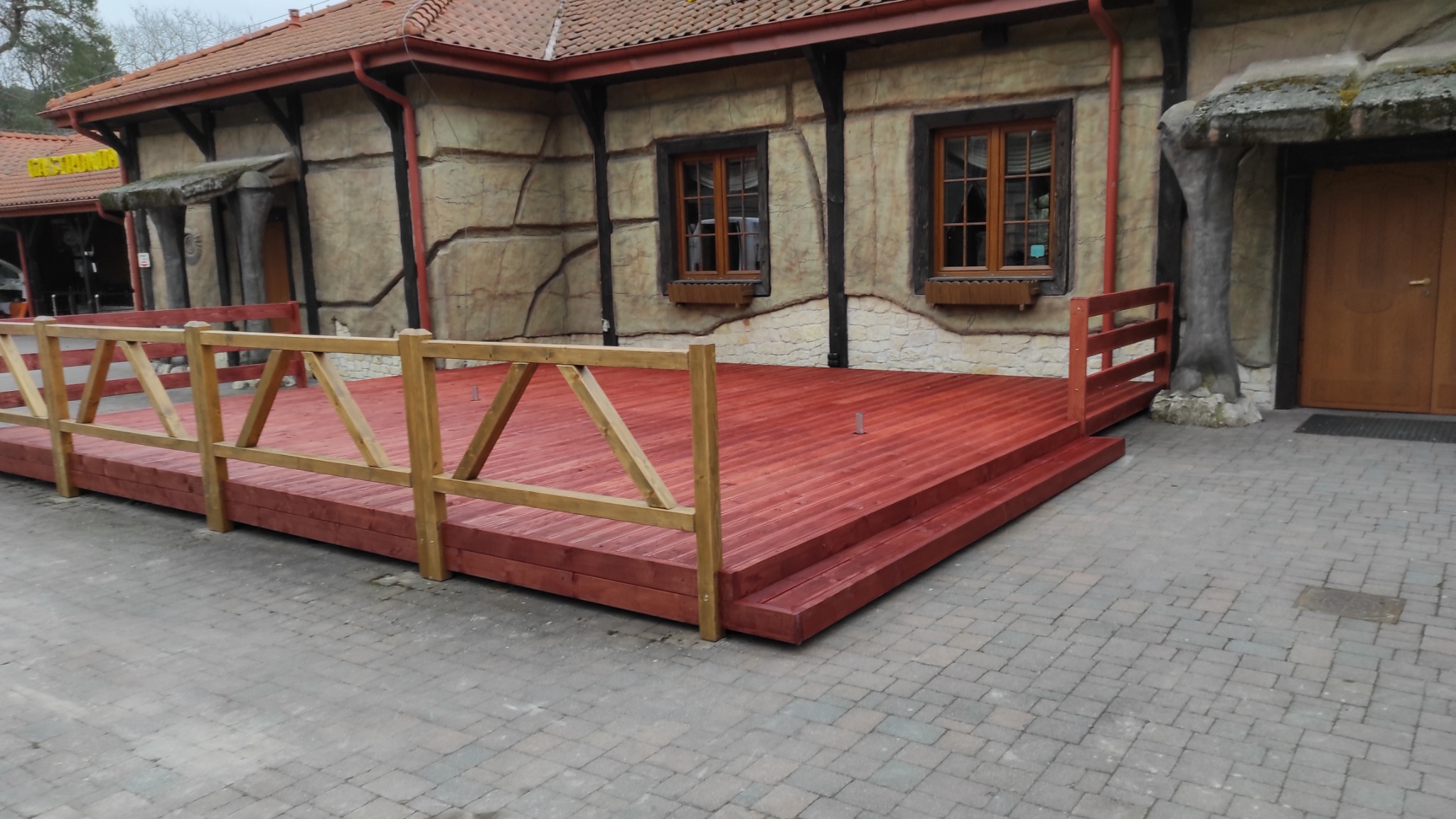 Czerwona drewniana platforma z balustradą przed budynkiem o elewacji imitującej kamień.