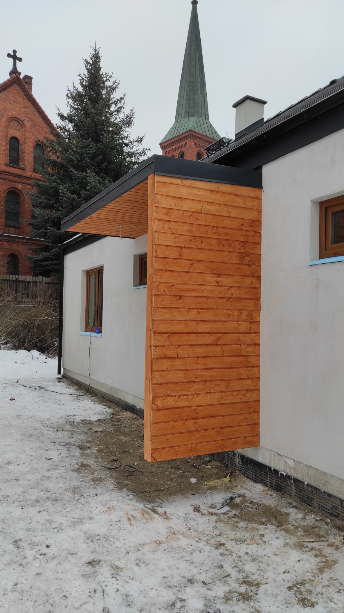Świeżo wykończona drewniana elewacja budynku w Solcu Kujawskim, z widoczną konstrukcją zadaszenia i fragmentem kościoła w tle, w zimowej scenerii.