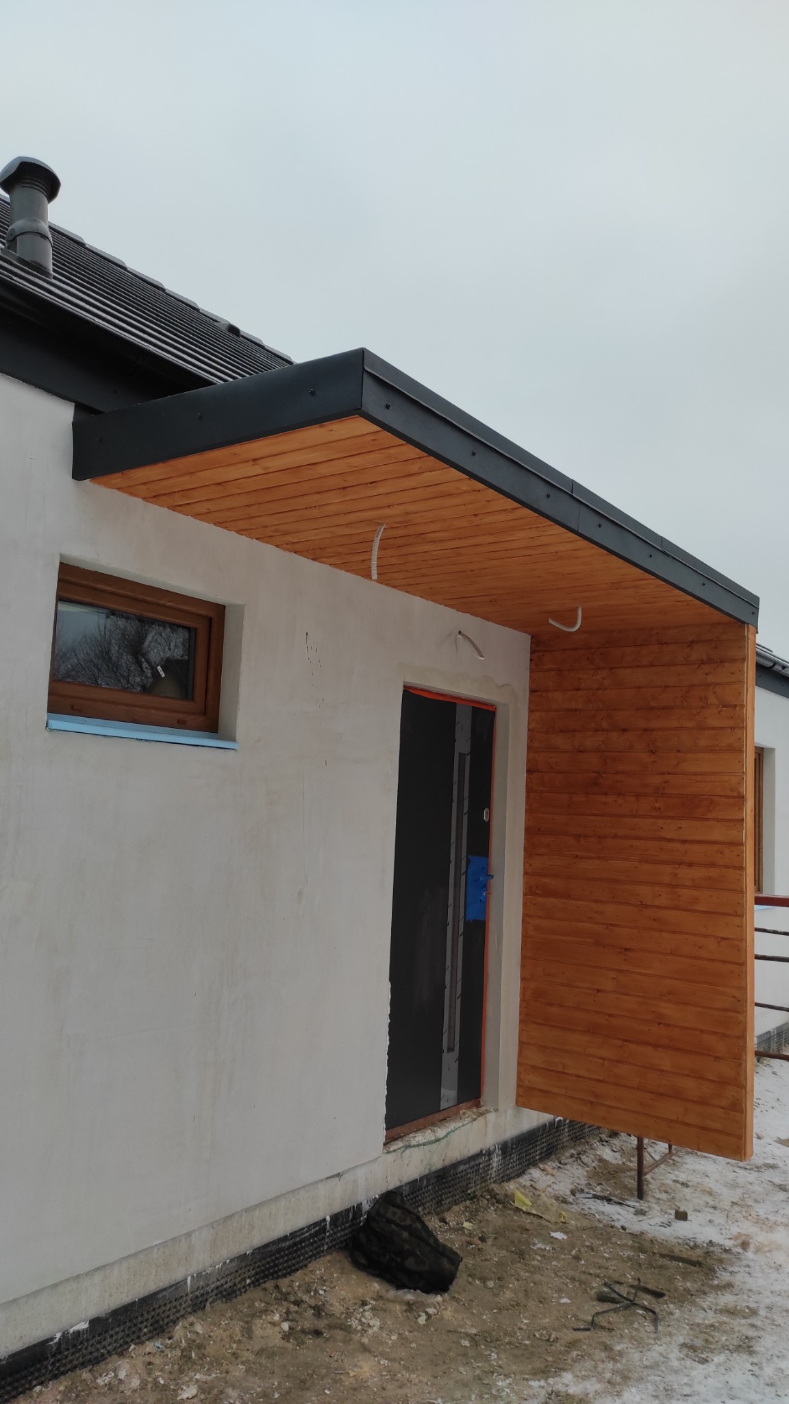 Surowy stan elewacji budynku z drewnianą wiatą nad wejściem, widoczne przewody elektryczne i zabezpieczenie przeciwwilgociowe fundamentów.