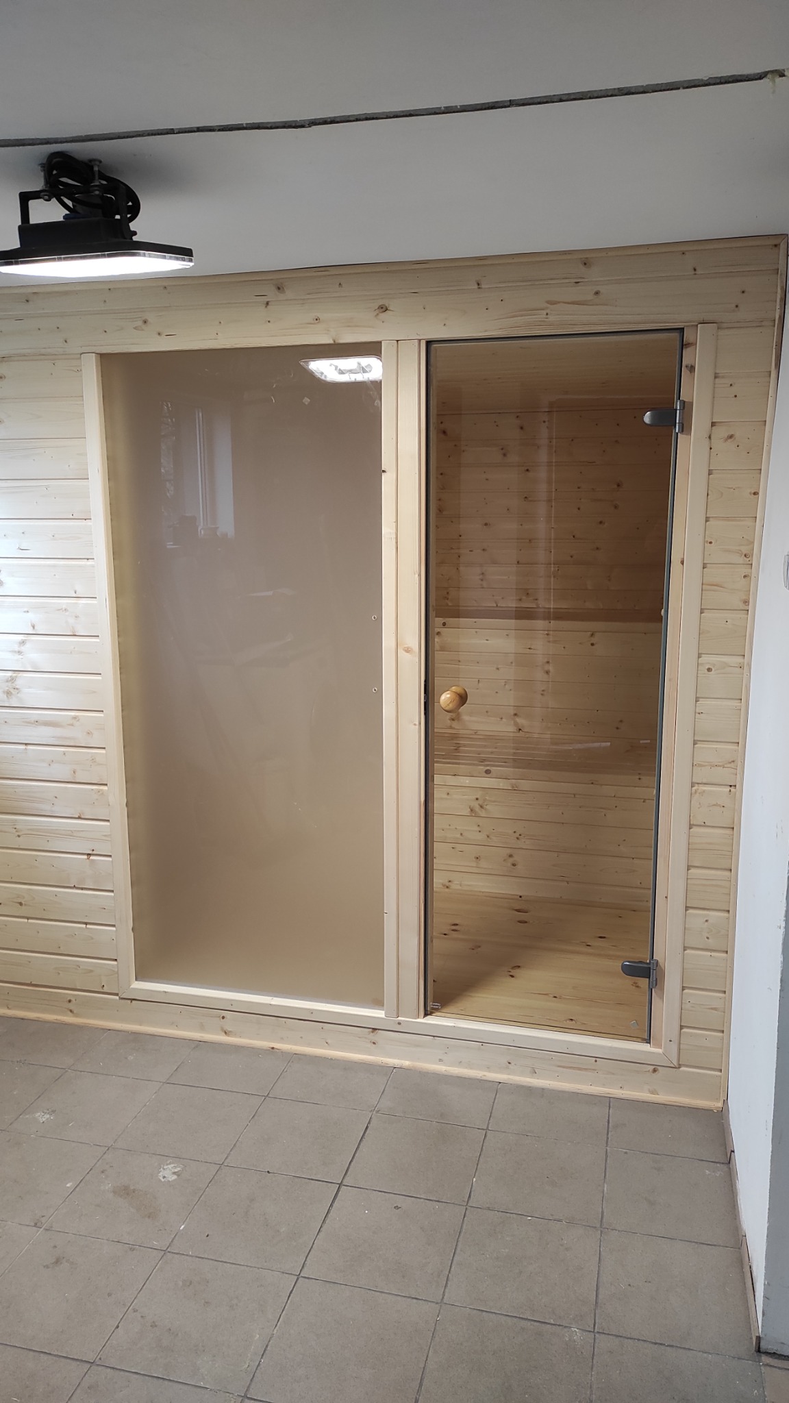 Wykończona sauna z drewnianą boazerią, szklanymi drzwiami z drewnianą gałką i matową szybą obok, widoczne szare płytki podłogowe.