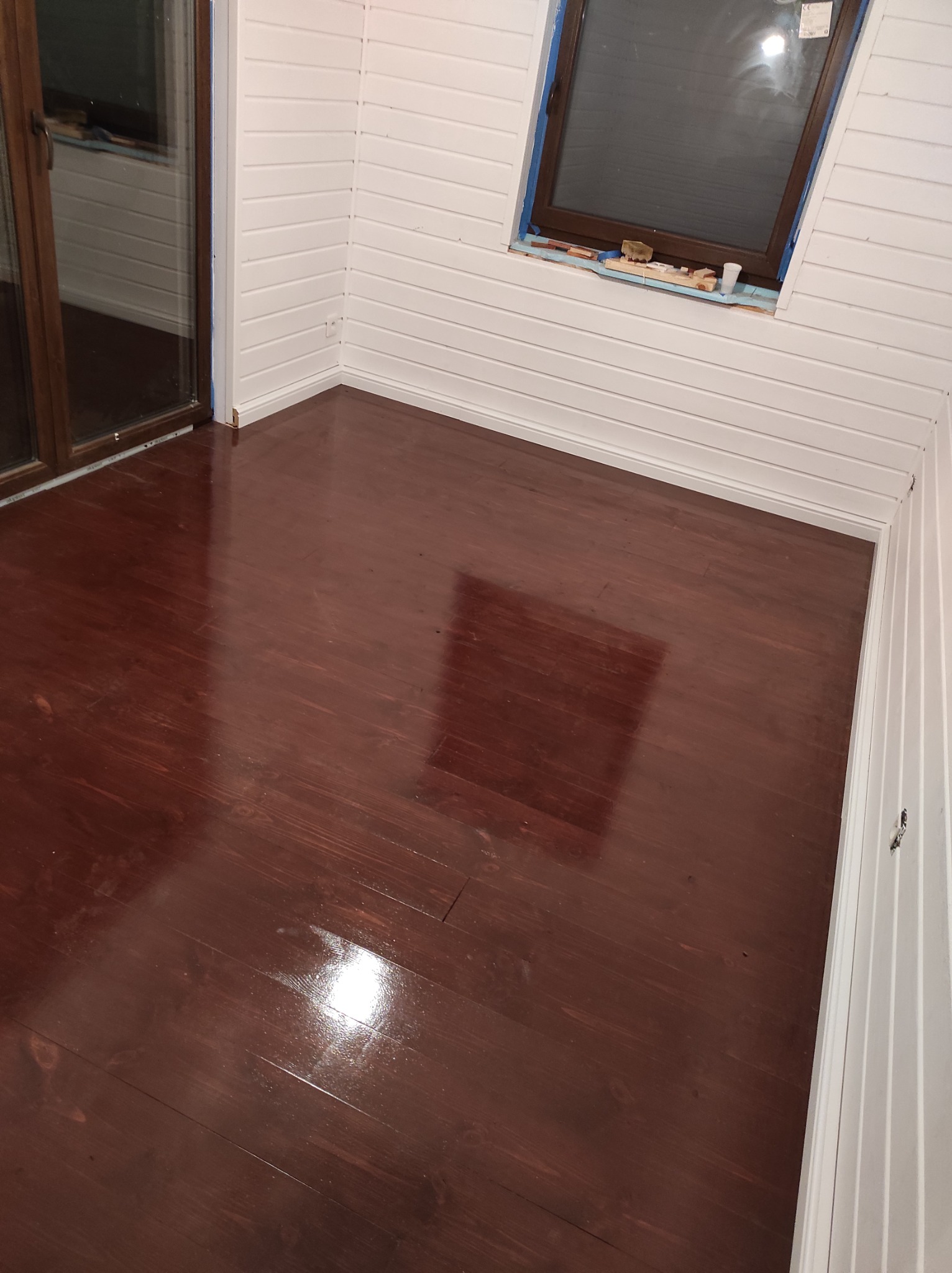 Świeżo polakierowana, ciemnobrązowa podłoga z desek w pokoju z białymi panelami ściennymi i oknem dachowym zabezpieczonym niebieską taśmą malarską.