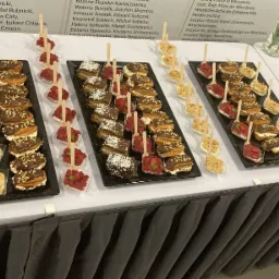 Elegancko podane przekąski na czarnych tackach: mini tartaletki z kremem, kostki ciasta z kokosem oraz mus owocowy na drewnianych łyżeczkach, ustawione na białym stole z czarną falbaną, w tle...