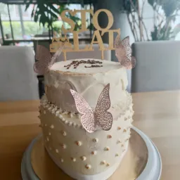 Dwupoziomowy tort urodzinowy w odcieniach bieli, udekorowany złotymi motylami i napisem 'STO LAT', umieszczony na złotym talerzu z szarą podstawą, w tle widoczne rośliny doniczkowe i krzesła.