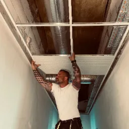 Mężczyzna z tatuażami na rękach montuje panel sufitowy w pomieszczeniu z widoczną instalacją wentylacyjną i kablową pod stropem.