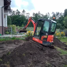 Czerwona mini koparka Kubota KX018-4 na gąsienicach stoi na stercie ziemi, w tle wykop pod fundamenty domu i ogród z żółtymi kwiatami.
