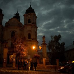 Fotografia Łukasz Kłoda - Grupa osób w eleganckich strojach stoi przed oświetlonym kościołem o zmierzchu, taksówki z żółtymi lampkami parkują w pobliżu.
