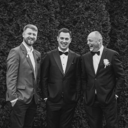 Fotografia Łukasz Kłoda - Trzech mężczyzn w garniturach i muchach, z uśmiechem pozujących na tle ciemnozielonego żywopłotu. Jeden z mężczyzn ma brodę i marynarkę w kratę, drugi w klasycznym smokingu, a trzeci śmieje się...