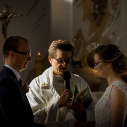 Fotografia Łukasz Kłoda - Uroczystość ślubna w kościele: ksiądz z mikrofonem błogosławi parę młodą, obrączki widoczne na okładce księgi.