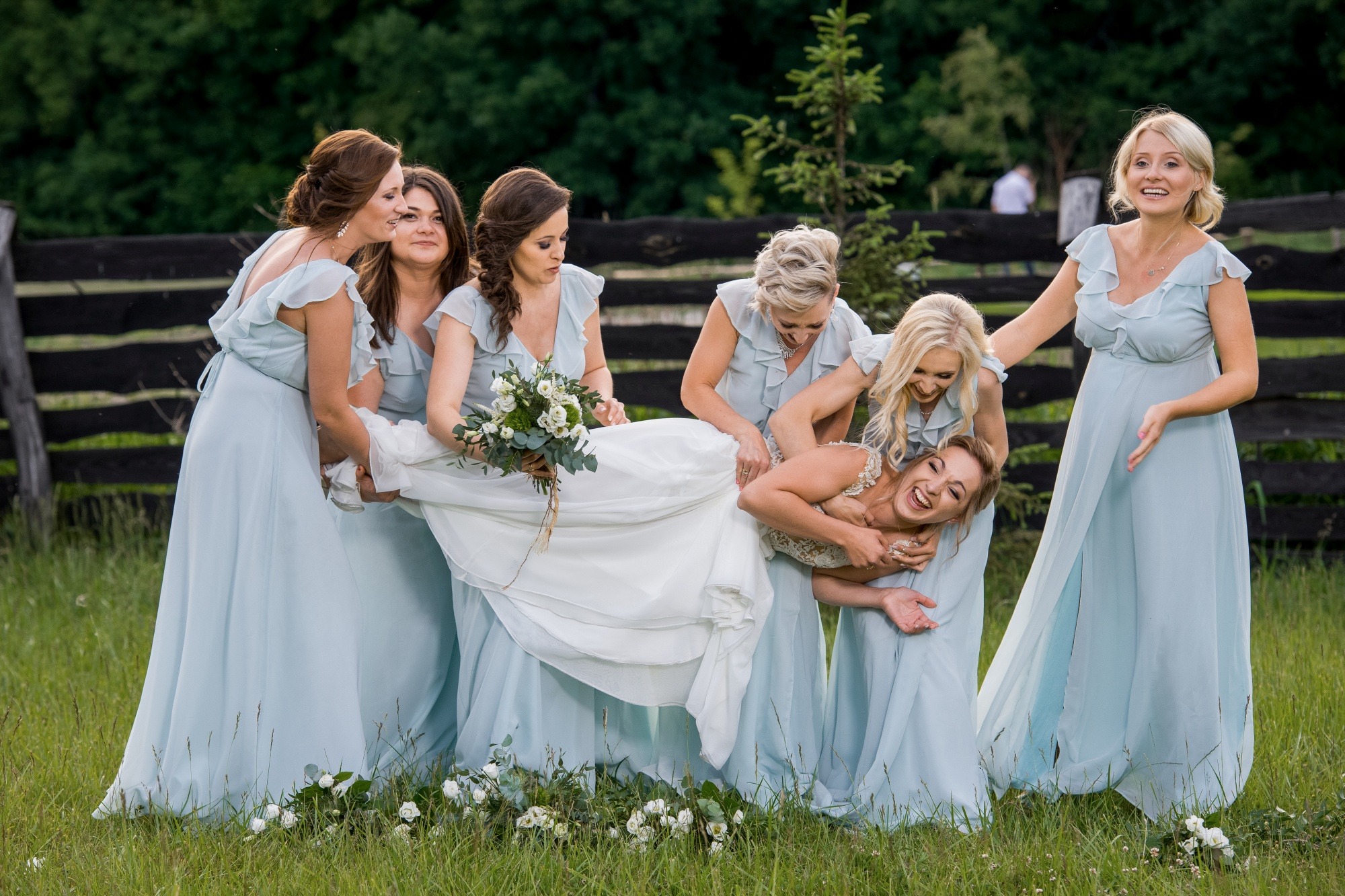 Panna młoda w białej sukni ślubnej, śmiejąca się, niesiona przez druhny w błękitnych sukienkach na tle zielonej trawy i drewnianego płotu.
