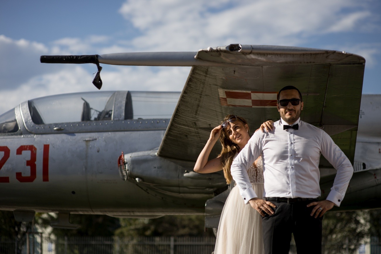 Para młoda pozuje przed srebrnym samolotem wojskowym z czerwonymi oznaczeniami, ona w sukni ślubnej i okularach na głowie, on w smokingu i ciemnych okularach, oboje uśmiechnięci.