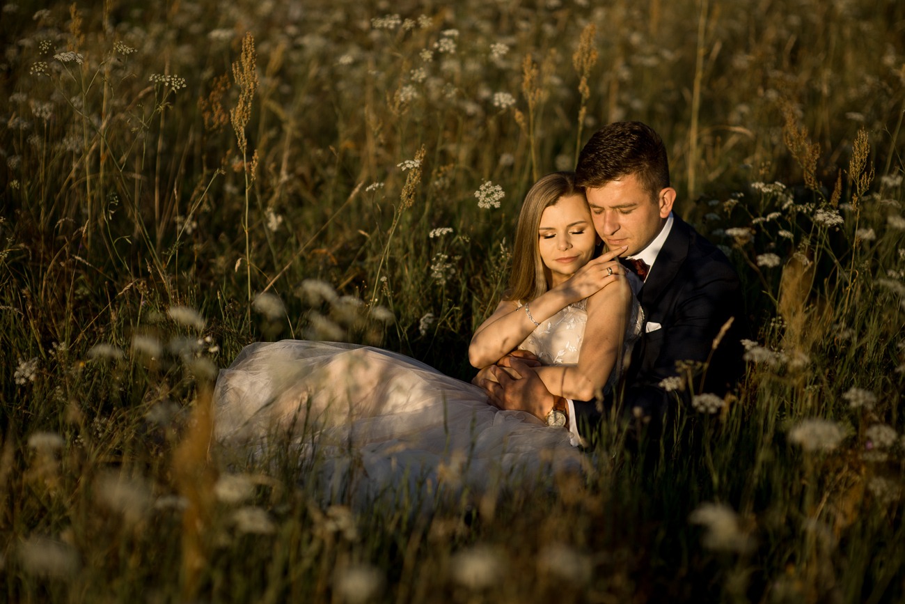 Para narzeczonych w objęciach siedzi na łące pełnej białych kwiatów i wysokich traw, delikatne światło podkreśla romantyczny nastrój.