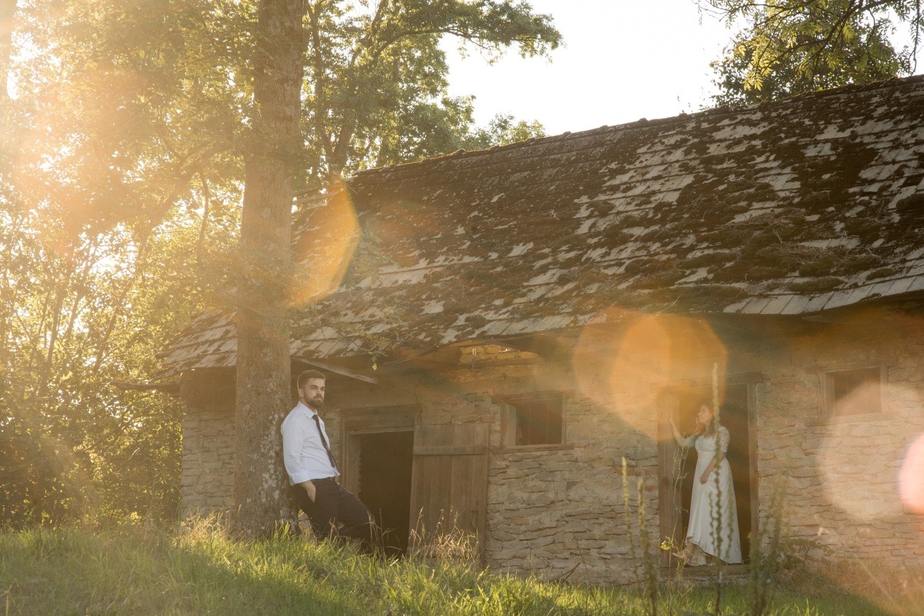 Para w rustykalnej scenerii: mężczyzna oparty o drzewo w koszuli i krawacie, kobieta w zwiewnej sukience w drzwiach kamiennego budynku z mchem na dachu, złote światło słoneczne.