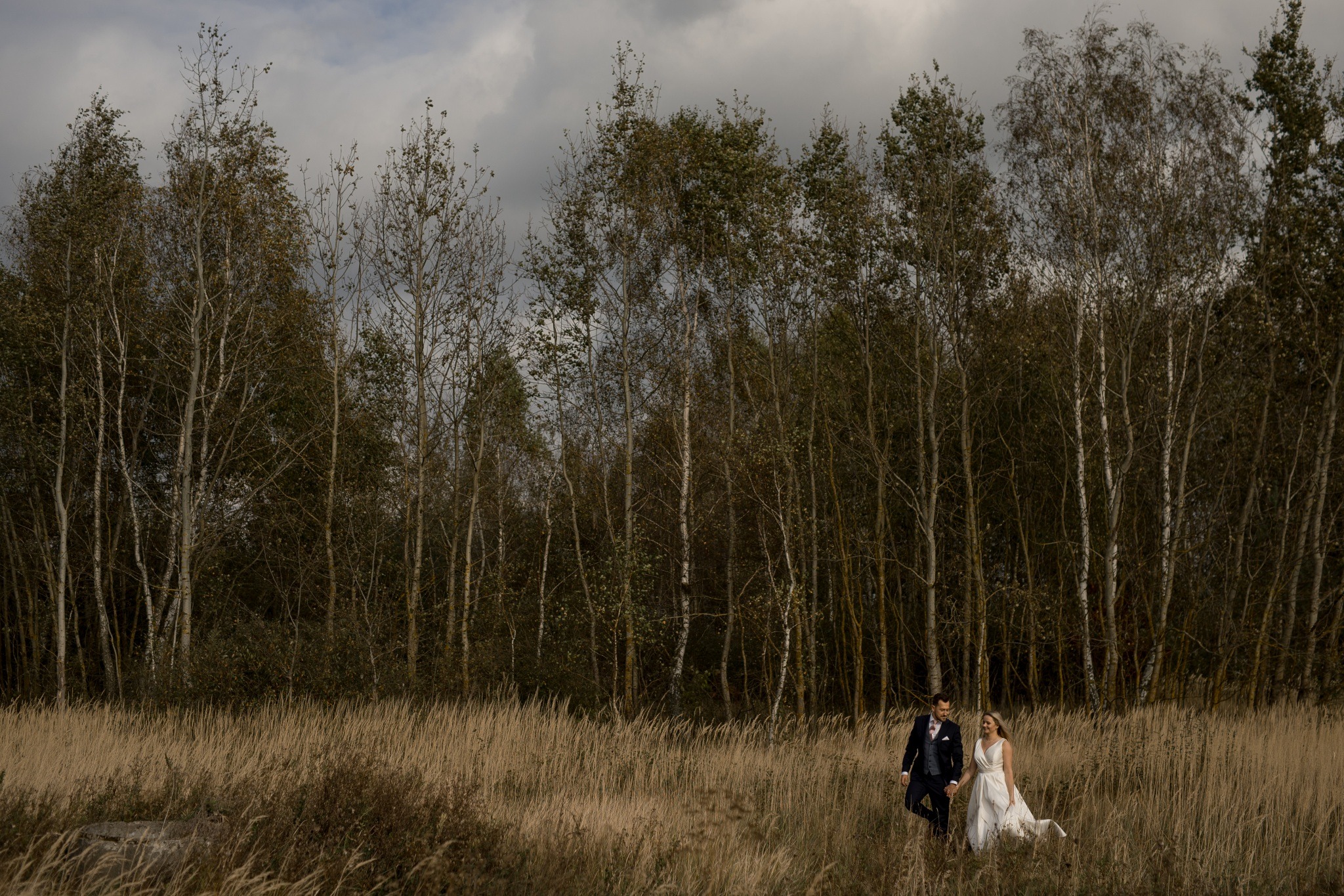 Para młoda idzie przez pole trzcin w kierunku lasu brzozowego pod pochmurnym niebem. Mężczyzna w granatowym garniturze i różową muszką, kobieta w białej sukni ślubnej z trenem.