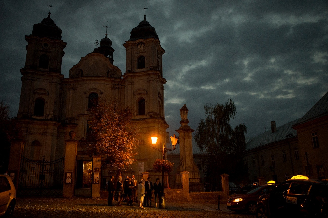 Grupa osób w eleganckich strojach stoi przed oświetlonym kościołem o zmierzchu, taksówki z żółtymi lampkami parkują w pobliżu.