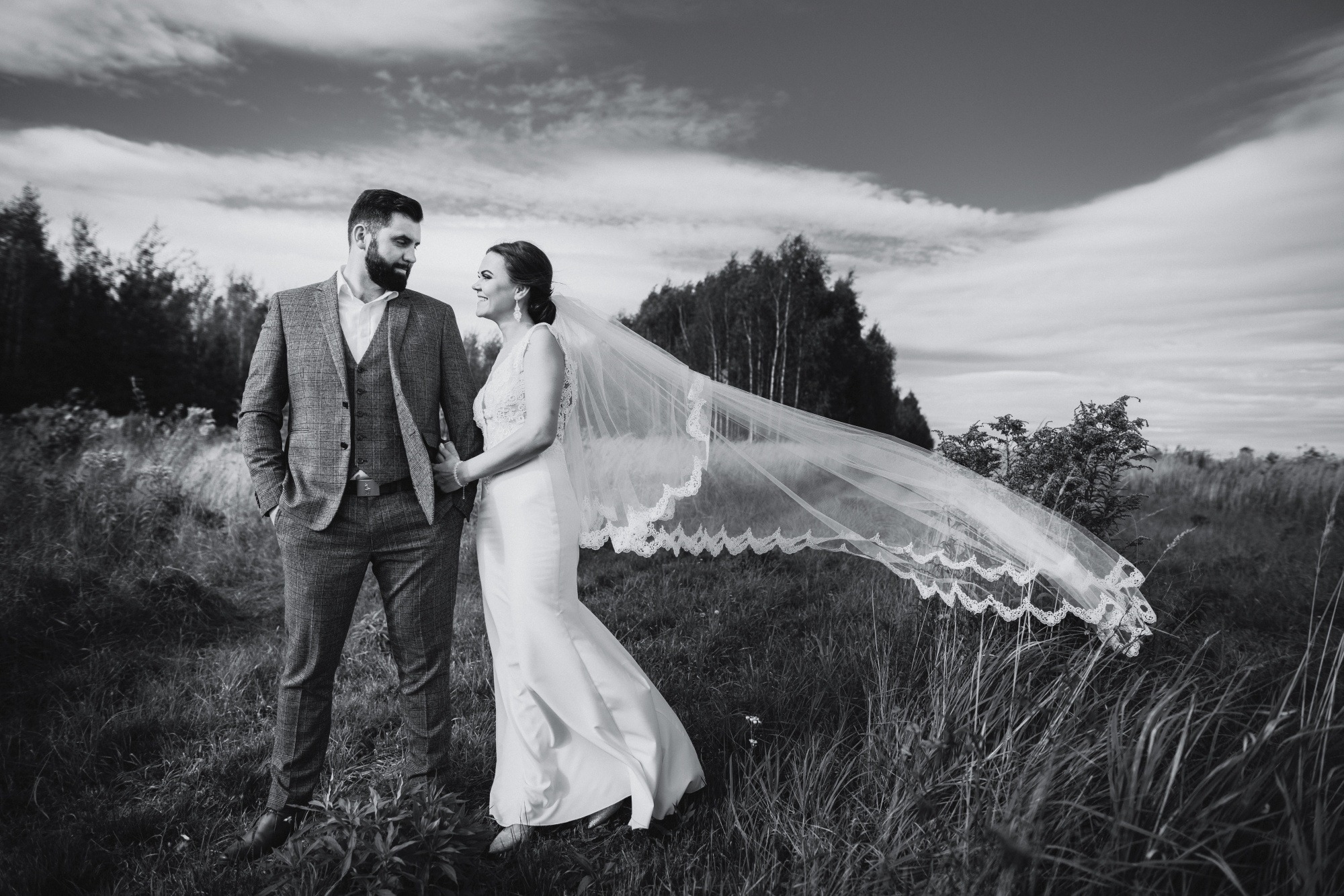 Czarno-białe zdjęcie narzeczonej w białej sukni z koronkowym wykończeniem welonu, który powiewa na wietrze, oraz narzeczonego w kraciastej, tweedowej marynarce i kamizelce, stojących na łące...