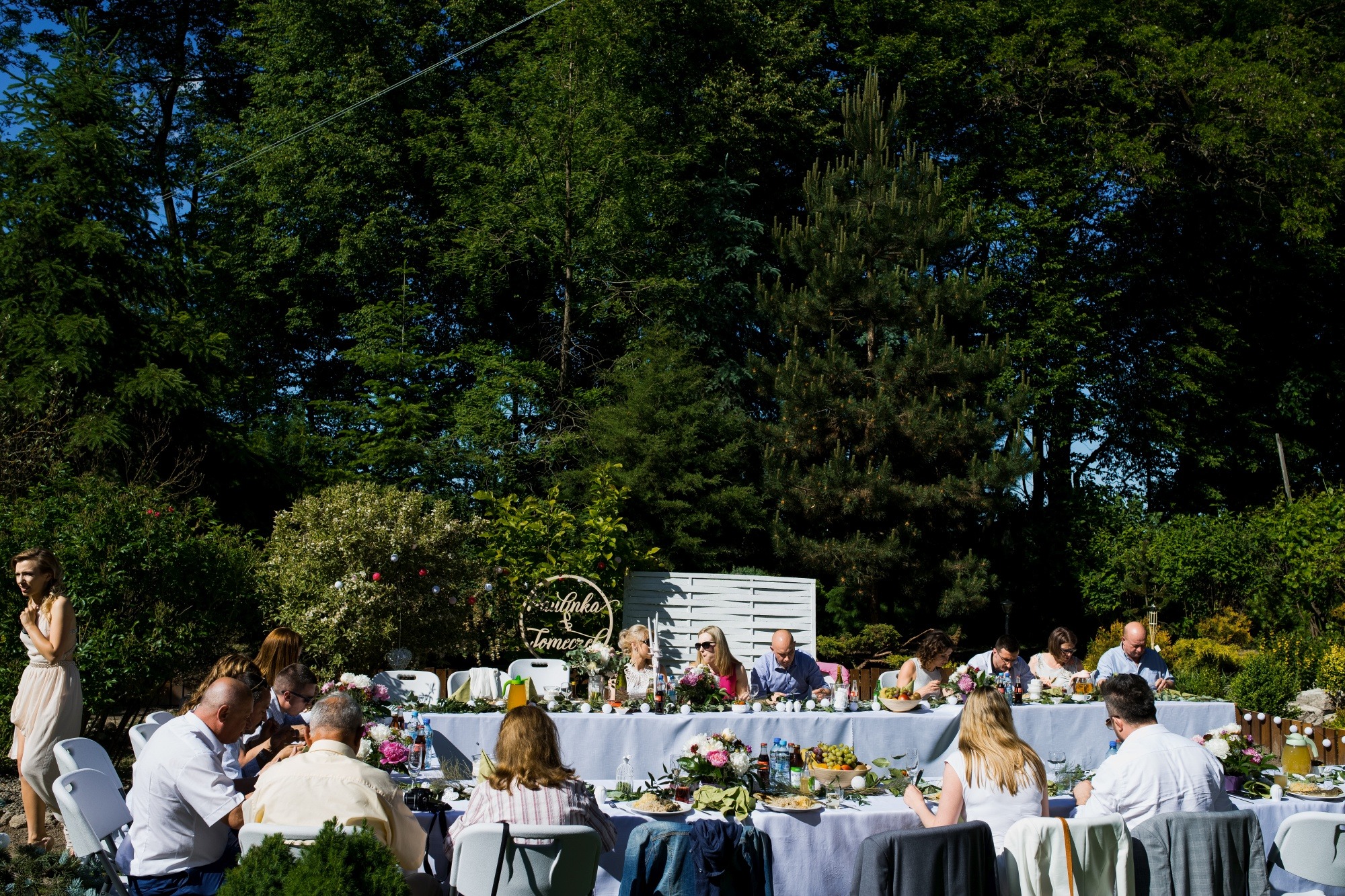 Długi stół z białym obrusem ustawiony na zewnątrz, otoczony gośćmi podczas uroczystości, dekoracje kwiatowe i girlandy świetlne, w tle zieleń drzew.