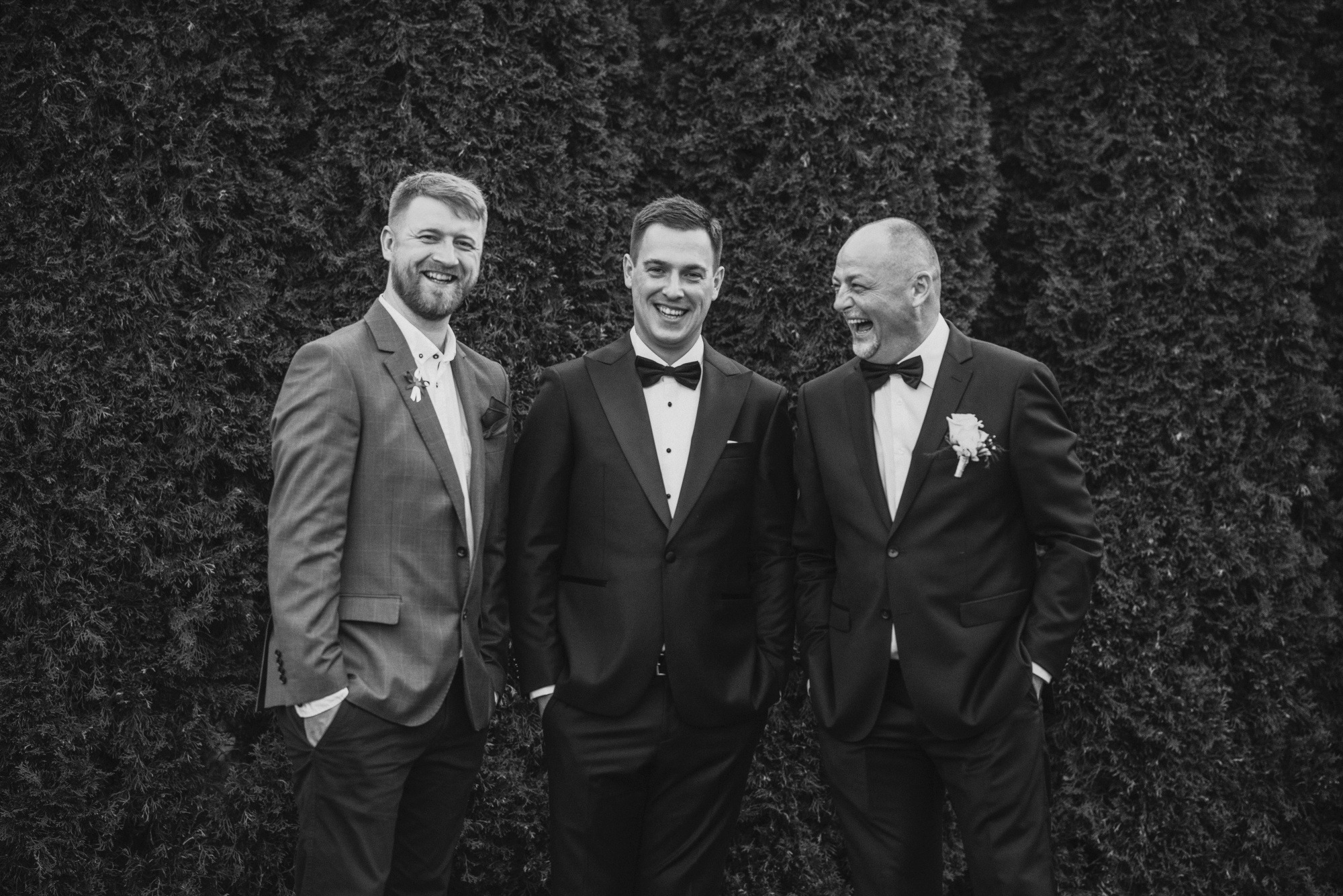 Trzech mężczyzn w garniturach i muchach, z uśmiechem pozujących na tle ciemnozielonego żywopłotu. Jeden z mężczyzn ma brodę i marynarkę w kratę, drugi w klasycznym smokingu, a trzeci śmieje się...