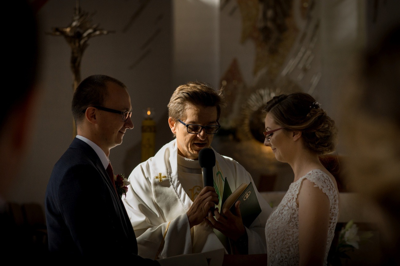 Uroczystość ślubna w kościele: ksiądz z mikrofonem błogosławi parę młodą, obrączki widoczne na okładce księgi.