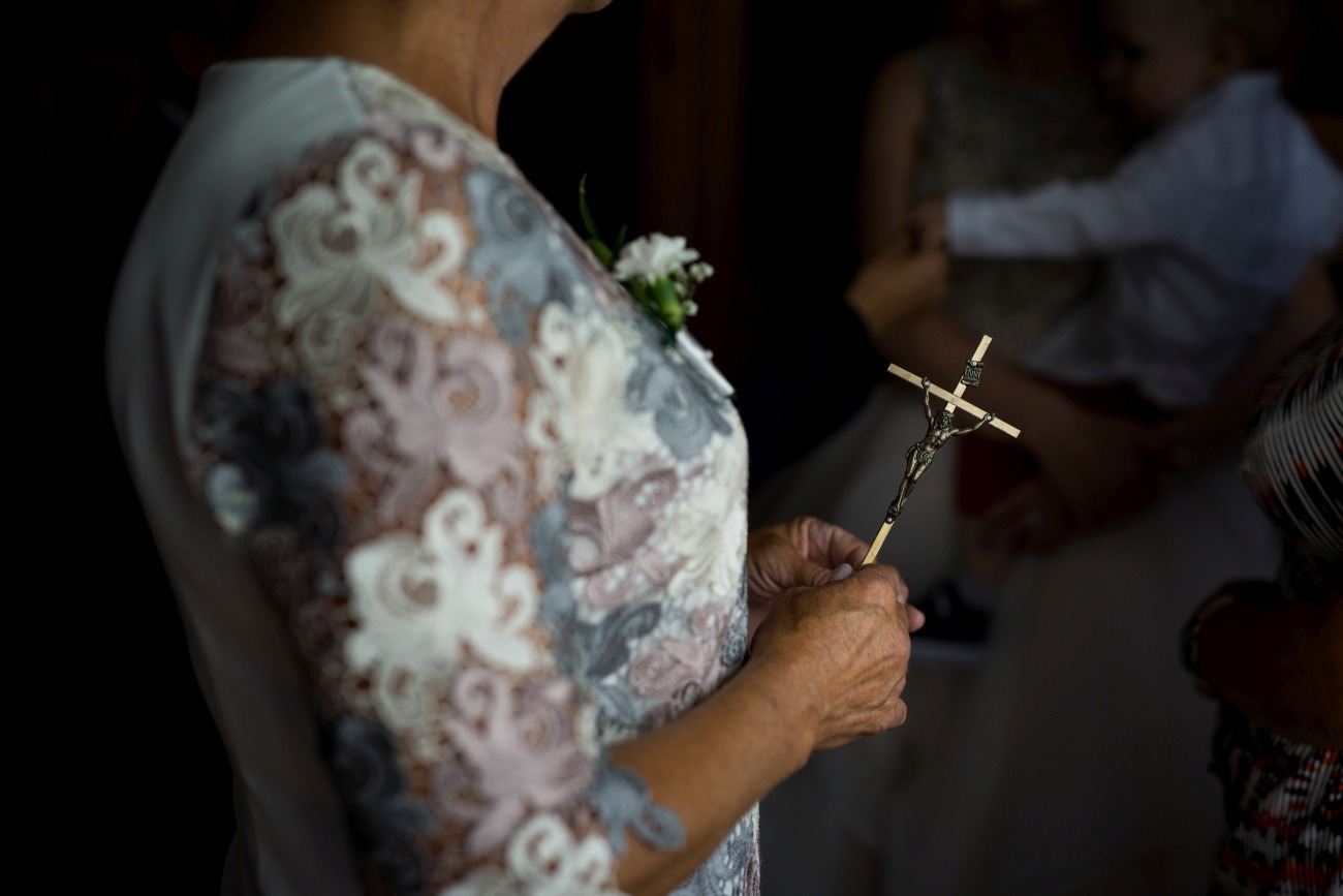 Kobieta w kwiecistej sukience trzyma w dłoniach krucyfiks na drewnianym patyczku, w tle niewyraźna postać dziecka na rękach.