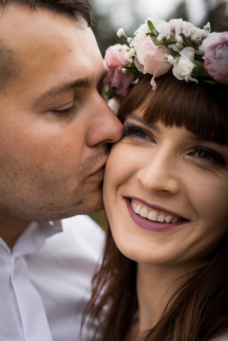 Uśmiechnięta kobieta w wianku z kwiatów, całowana w policzek przez mężczyznę. Makro zbliżenie na twarz, widoczne sztuczne rzęsy i matowa pomadka.