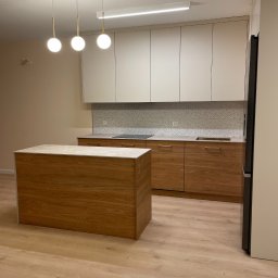 STOLARSTWO MEBLOWE JADWIGA SZAFLARSKA - Nowoczesna kuchnia z drewnianą wyspą i białymi szafkami górnymi. Nad wyspą designerska lampa z trzema kulami. Jasna podłoga i mozaika nad blatem dodają elegancji.