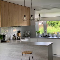 STOLARSTWO MEBLOWE JADWIGA SZAFLARSKA - Nowoczesna kuchnia z drewnianymi szafkami wiszącymi, szarym blatem i wyspą. Nad wyspą wiszą trzy lampy z geometrycznymi kloszami. Za oknem zieleń.