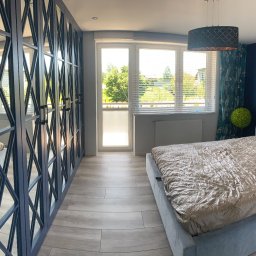 STOLARSTWO MEBLOWE JADWIGA SZAFLARSKA - Szafa wnękowa z lustrami w niebieskiej ramie, z geometrycznym wzorem, w jasnej sypialni z łóżkiem i oknem balkonowym. Widok z perspektywy.