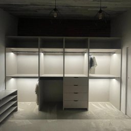 STOLARSTWO MEBLOWE JADWIGA SZAFLARSKA - Jasna szafa wnękowa z oświetleniem LED, widok z przodu. Ubrania wiszą na wieszakach. Na podłodze folia ochronna. Dwie lampy wiszące na suficie.