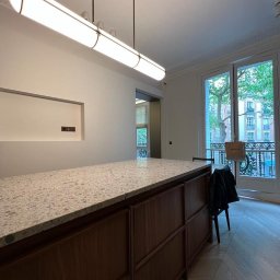 oOomeble.pl - Nowoczesna wyspa kuchenna z blatem lastryko i drewnianymi frontami, oświetlona podłużną lampą. Widok na balkon przez okno.