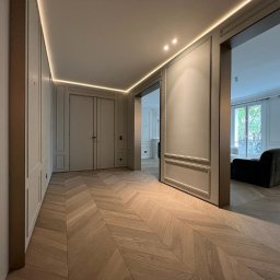 STOLARSTWO MEBLOWE JADWIGA SZAFLARSKA - Eleganckie wnętrze z parkietem w jodełkę, listwami ściennymi i subtelnym oświetleniem LED. Widok na salon z sofą i oknami balkonowymi.