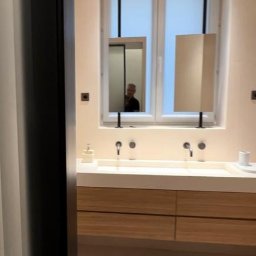 STOLARSTWO MEBLOWE JADWIGA SZAFLARSKA - Nowoczesna łazienka z drewnianą szafką pod umywalką i oryginalnymi lustrami wiszącymi na czarnych prętach. W tle okno z roletami.