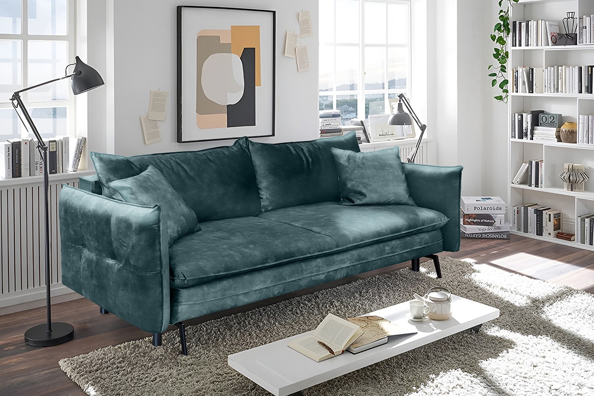 Stylowa, turkusowa sofa z poduszkami w salonie. Na stoliku książka i serwis do herbaty. W tle regał z książkami, okno i lampa.