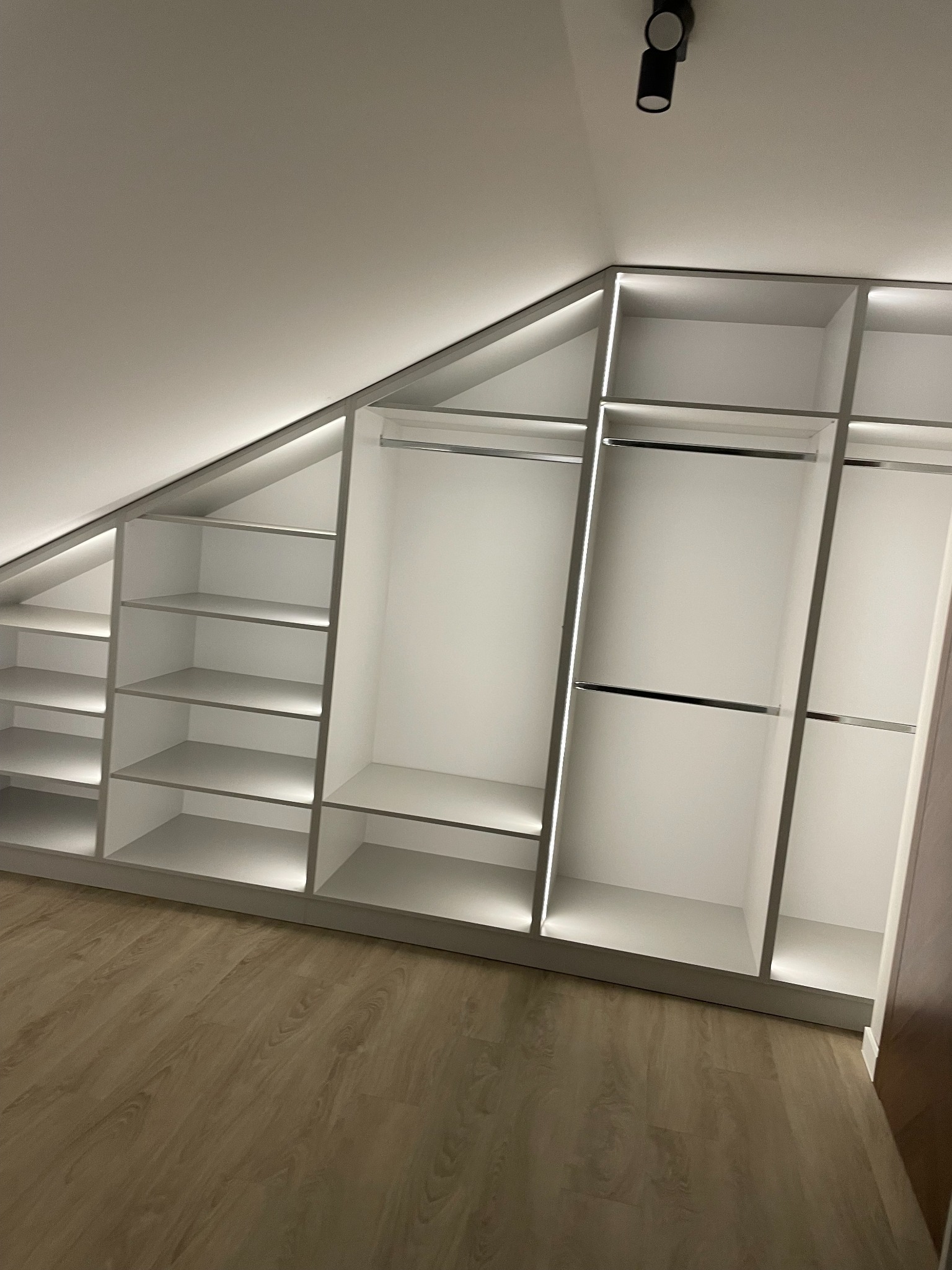 Biała szafa wnękowa na poddaszu z oświetleniem LED, półki i drążki na ubrania, drewniana podłoga, minimalistyczny design, czarny reflektor sufitowy.