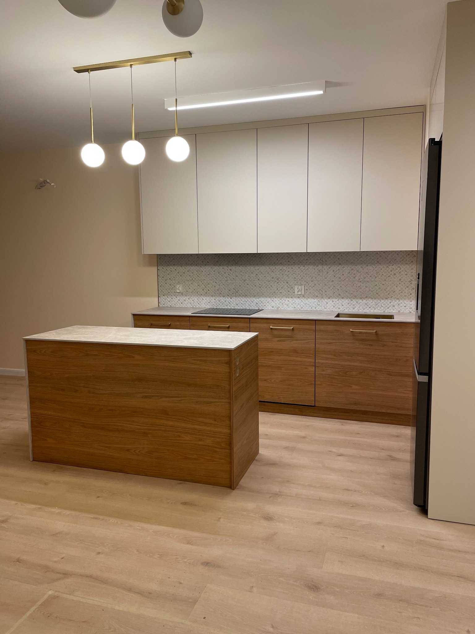 Nowoczesna kuchnia z drewnianą wyspą i białymi szafkami górnymi. Nad wyspą designerska lampa z trzema kulami. Jasna podłoga i mozaika nad blatem dodają elegancji.