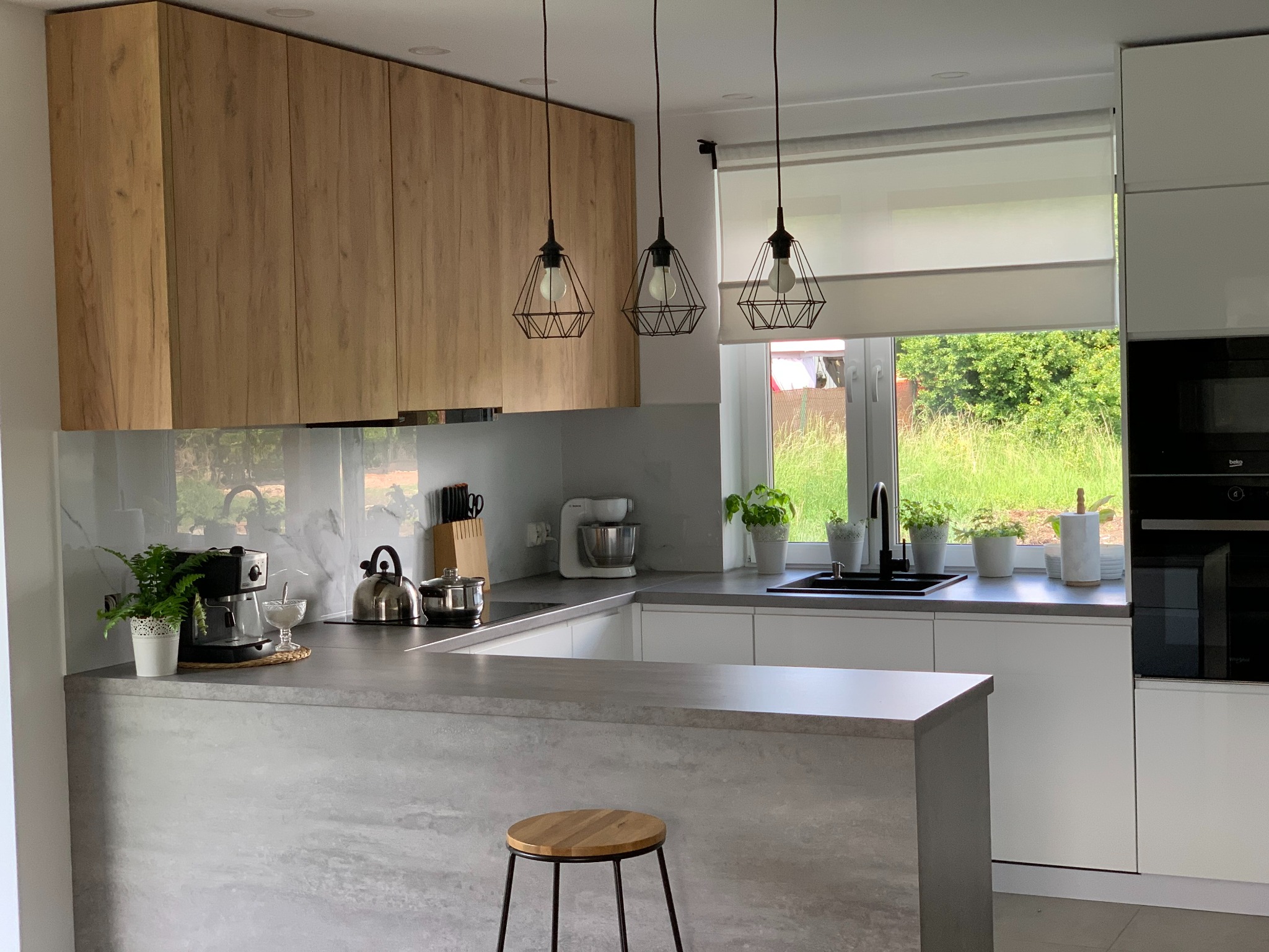 Nowoczesna kuchnia z drewnianymi szafkami wiszącymi, szarym blatem i wyspą. Nad wyspą wiszą trzy lampy z geometrycznymi kloszami. Za oknem zieleń.
