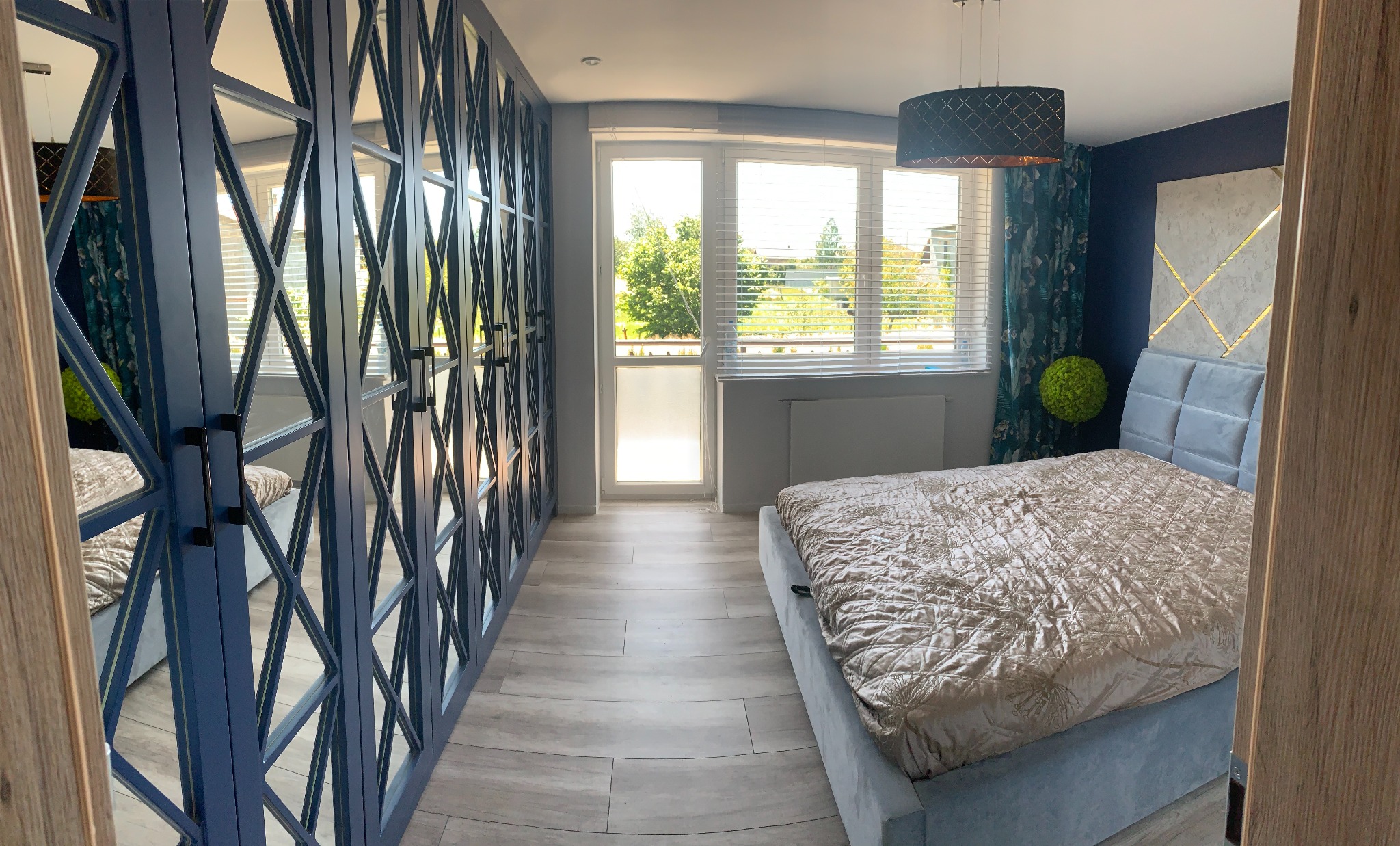 Szafa wnękowa z lustrami w niebieskiej ramie, z geometrycznym wzorem, w jasnej sypialni z łóżkiem i oknem balkonowym. Widok z perspektywy.