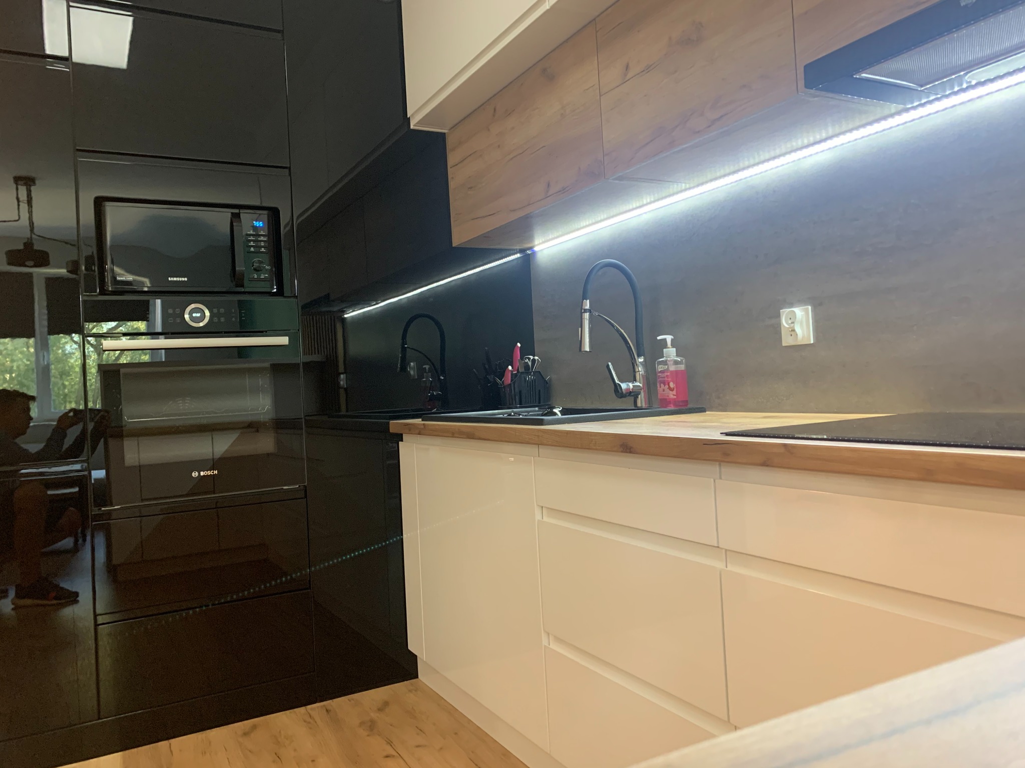 Nowoczesna kuchnia z czarnymi, błyszczącymi szafkami i białymi frontami dolnymi. Drewniany blat, oświetlenie LED pod szafkami górnymi. Widoczny sprzęt AGD.