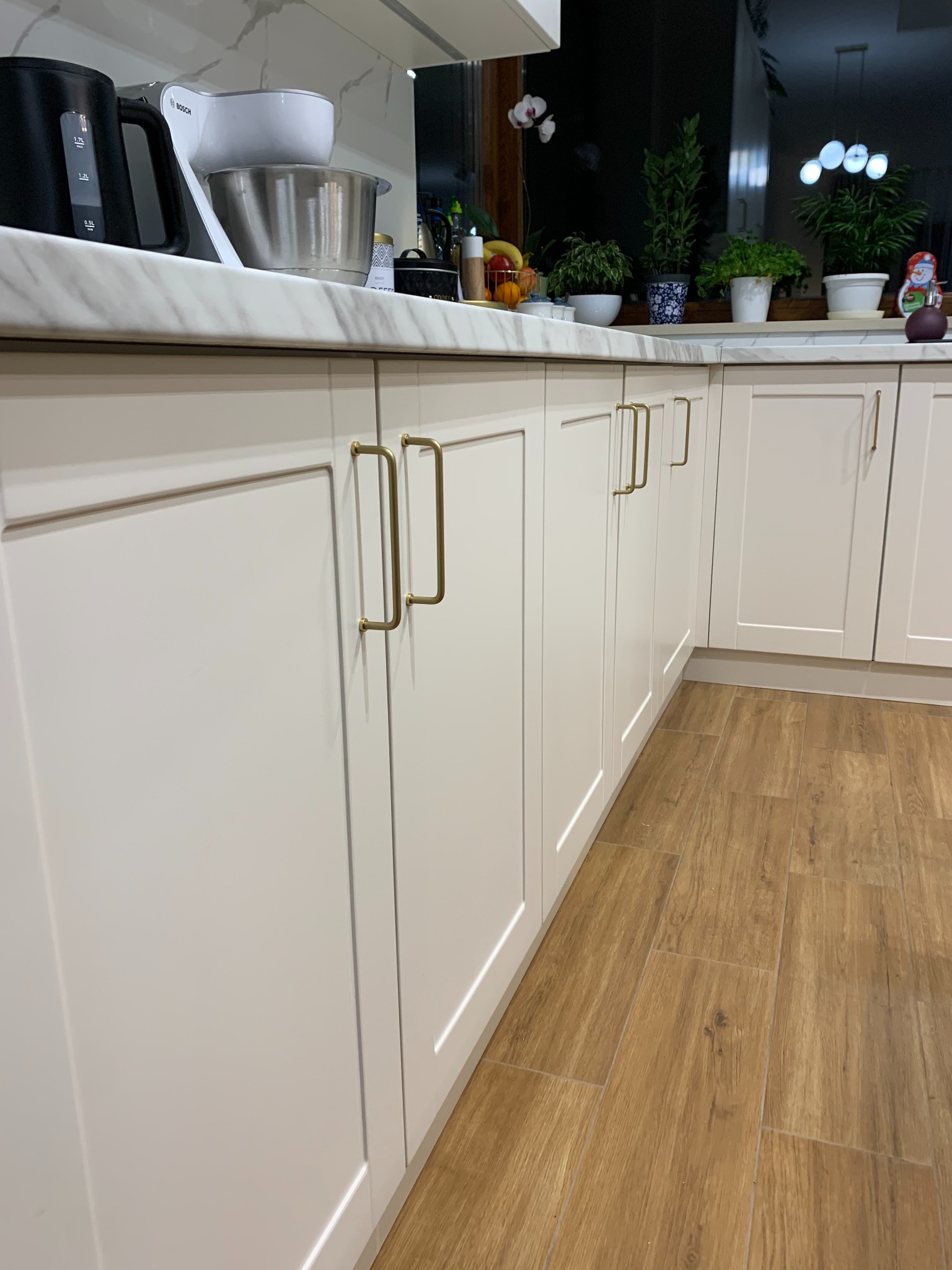 Jasne, frezowane fronty szafek kuchennych z podłużnymi, złotymi uchwytami. Na blacie marmurowy wzór, a w tle okno z roślinami doniczkowymi.