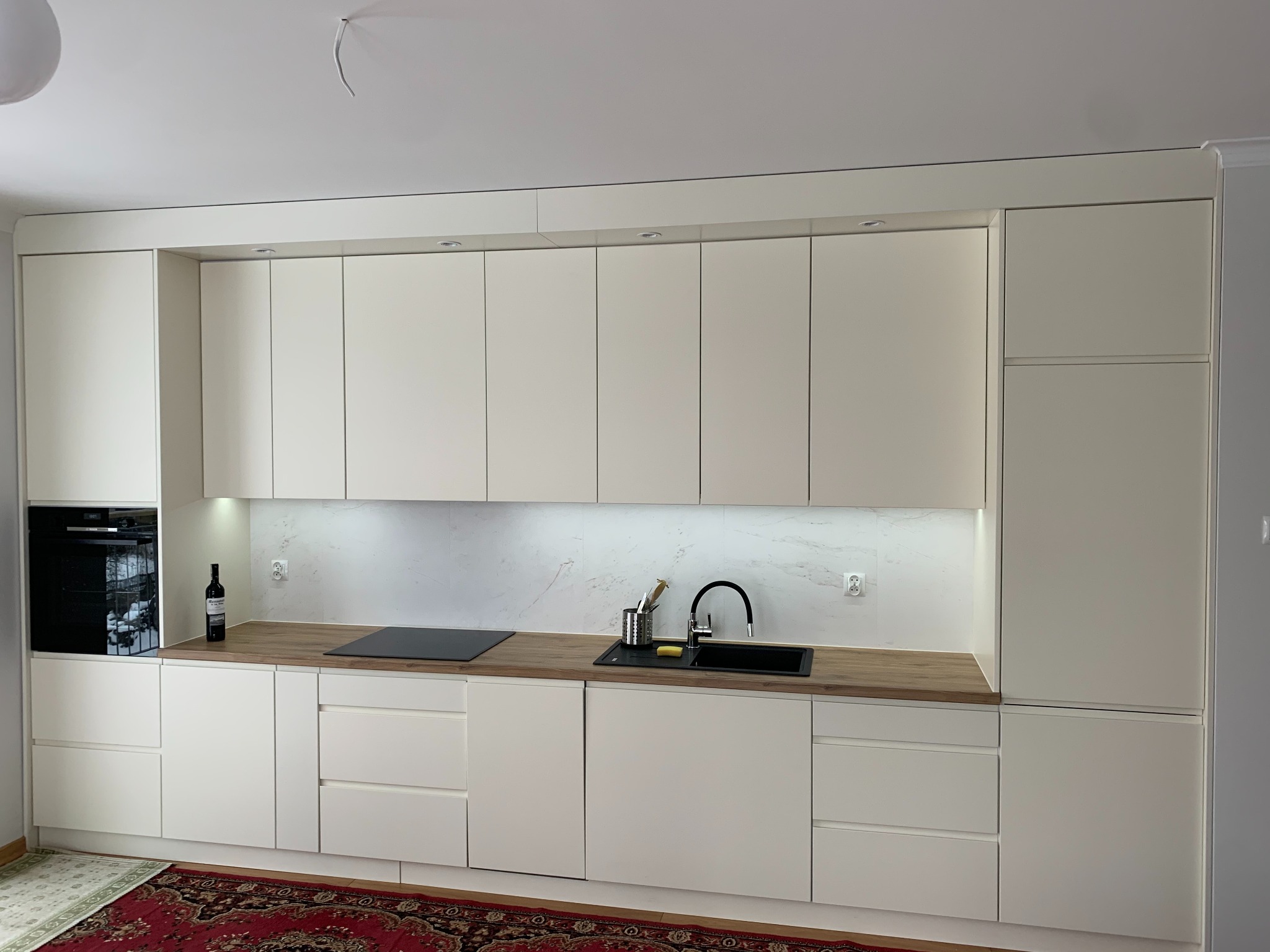 Jasna kuchnia z drewnianym blatem i marmurowym panelem. Uwagę zwraca czarna armatura i butelka wina obok piekarnika. Całość oświetlona punktowo.