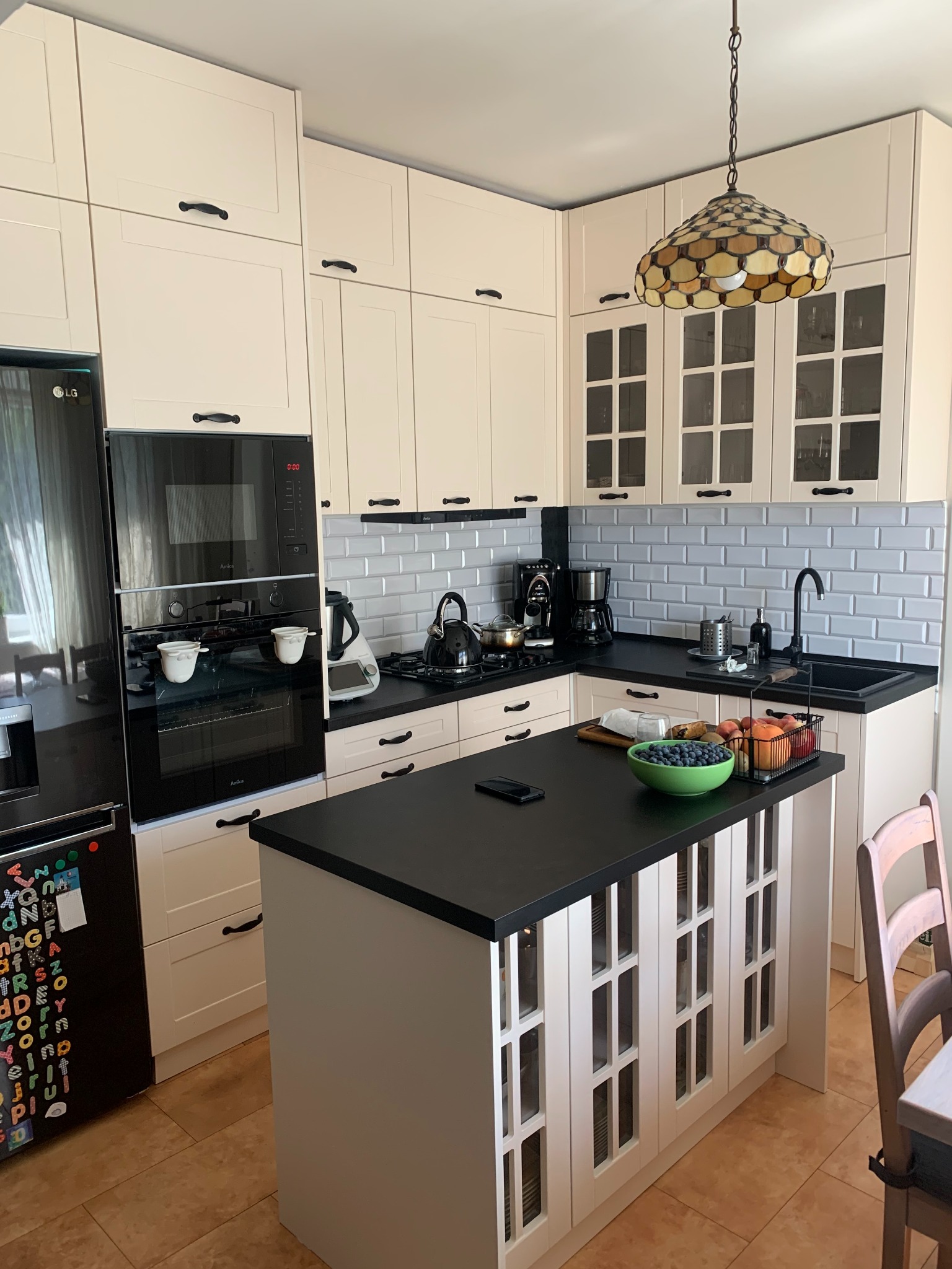 Jasna kuchnia z wyspą i czarnym blatem. Na wyspie owoce w misce. Nad wyspą wisi lampa w stylu Tiffany. Białe szafki z czarnymi uchwytami. Na lodówce magnesy.