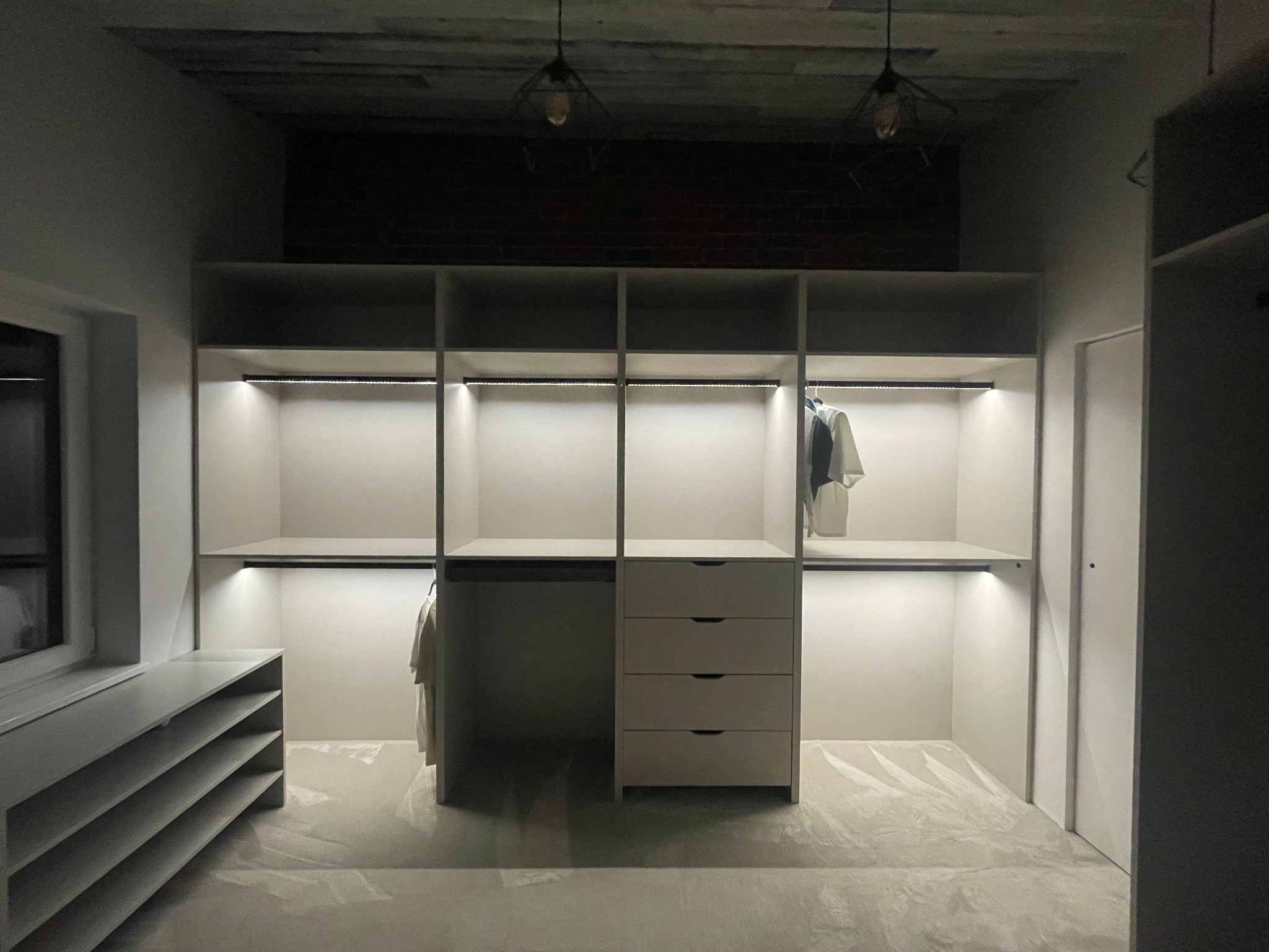 Jasna szafa wnękowa z oświetleniem LED, widok z przodu. Ubrania wiszą na wieszakach. Na podłodze folia ochronna. Dwie lampy wiszące na suficie.