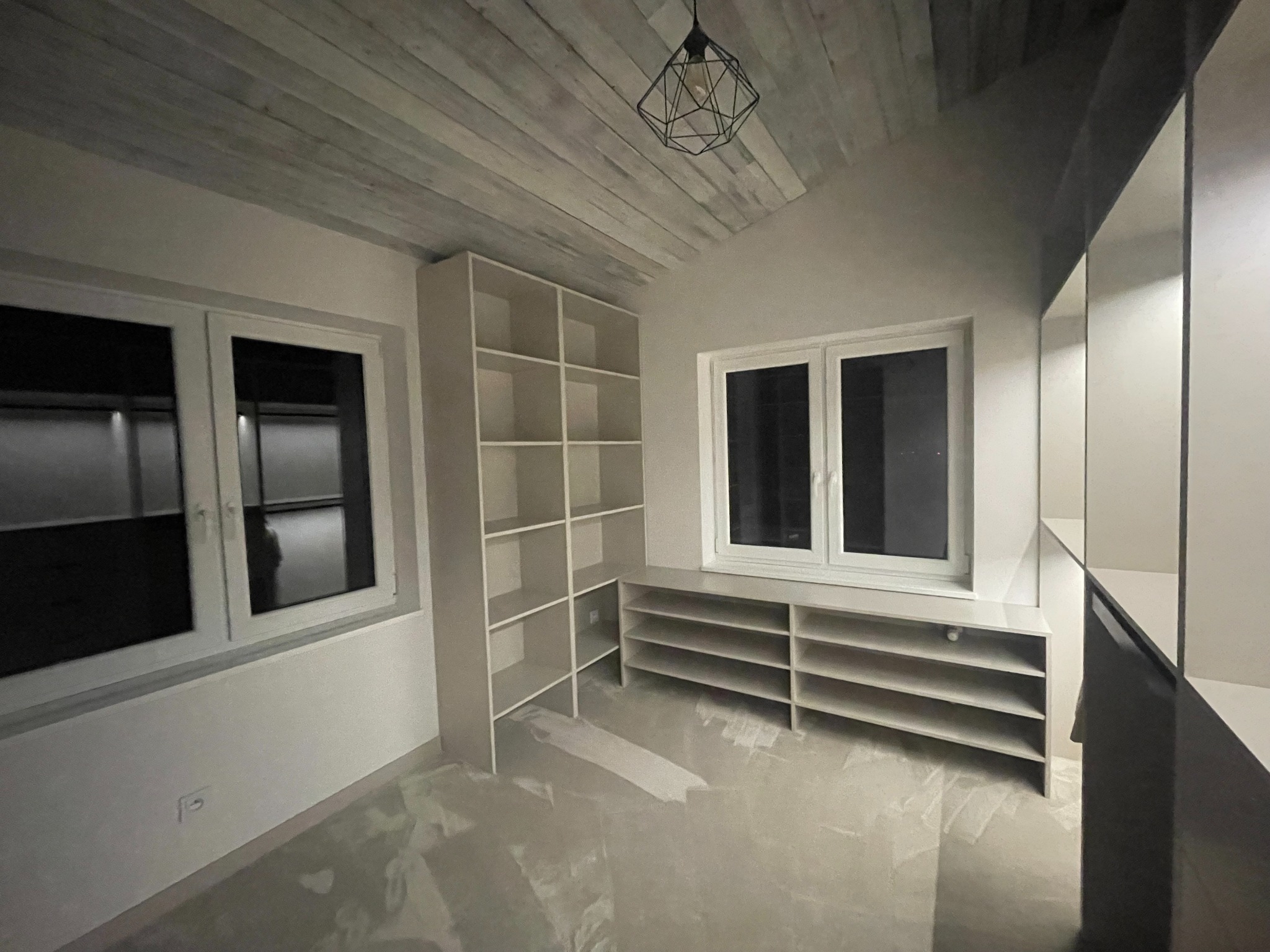 Jasne, minimalistyczne wnętrze z regałami i szafkami pod oknem. Widoczny sufit z drewnianych desek i nowoczesna lampa. Podłoga zabezpieczona folią malarską.