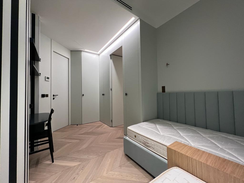 Nowoczesny pokój z łóżkiem z tapicerowanym zagłówkiem, biurkiem i szafami w zabudowie. Parkiet w jodełkę, minimalistyczne oświetlenie LED.